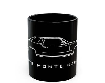 May include: A black ceramic mug featuring a white line art illustration of a 1973 Monte Carlo car on the side. Below the car graphic, the text 1973 MONTE CARLO is printed in a clean, white, sans-serif font against the solid black background.