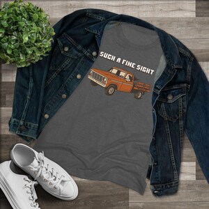 May include: A gray t-shirt with the phrase "SUCH A FINE SIGHT" above an orange pickup truck graphic. The shirt is displayed with a denim jacket and white sneakers on a wooden surface.