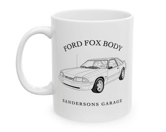 May include: White ceramic mug featuring a black line drawing of a Ford Fox Body car. The text "FORD FOX BODY" is above the car image, and "SANDERSONS GARAGE" is below. The mug has a curved handle.