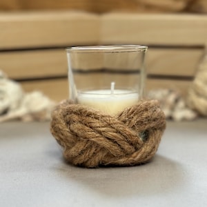 May include: A clear glass votive candle holder with a white candle inside, encircled by a decorative brown rope knot. The candle holder sits on a gray surface, with a wooden background.
