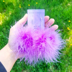 May include: Lavender feather earrings with silver chain detailing. The earrings are presented on a card that reads "KILVER KAT CREATIONS STASH EARRINGS". The background is a blurred green field.