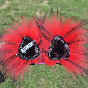 May include: A pair of red and black faux fur hair clips with black sequins and a silver bar with the word "RIDDIM" on it.