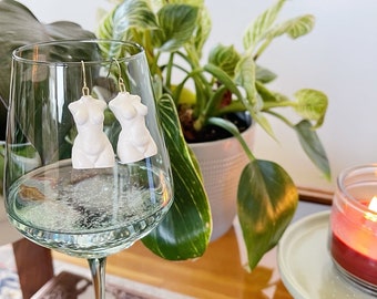 May include: White ceramic earrings shaped like the human torso, hanging from a glass of white wine. The earrings are on a gold hook.