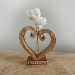 May include: Wooden heart-shaped vase with a glass tube for flowers. The vase has a carved design and the words "Love is in the air..." are engraved on the base.