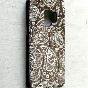 May include: A brown phone case with a white paisley pattern. The case is made of wood and has a black border.