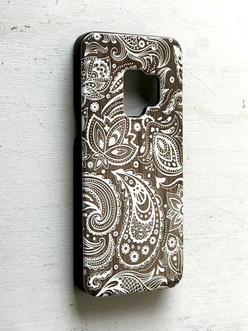 May include: A brown phone case with a white paisley pattern. The case is made of wood and has a black border.