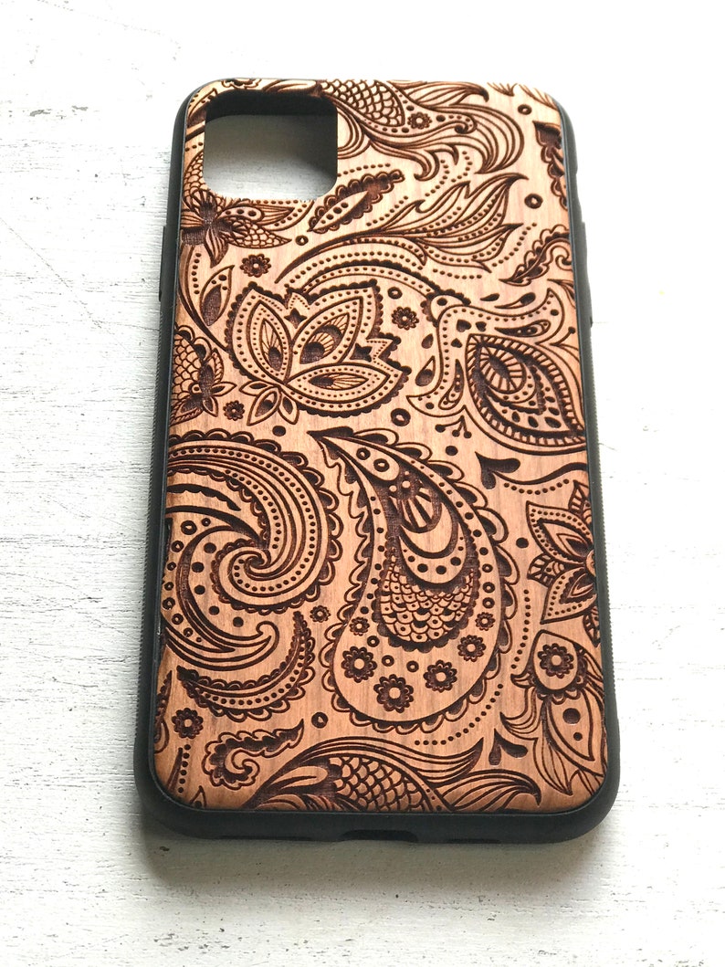 May include: A wooden phone case with a paisley pattern carved into the surface. The case has a black border.