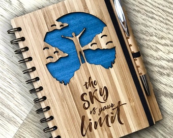 Puede incluir: Un diario de madera con encuadernación en espiral y un bolígrafo. La portada presenta un cielo azul con nubes y una silueta de una persona con los brazos extendidos. El texto "the sky is your limit" está grabado en la portada.