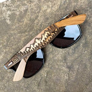 May include: Wooden sunglasses with black lenses and a mountain and forest design on the arms. The arms are engraved with the text "Stay Wild".