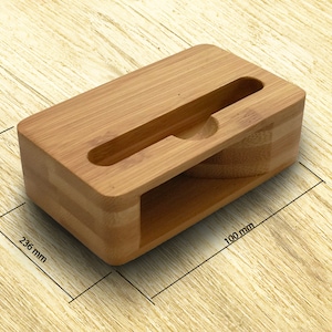 Może przedstawiać: Stojak na telefon z naturalnego bambusa z wycięciem na telefon. Stojak ma wymiary 100 mm x 236 mm x 30 mm.