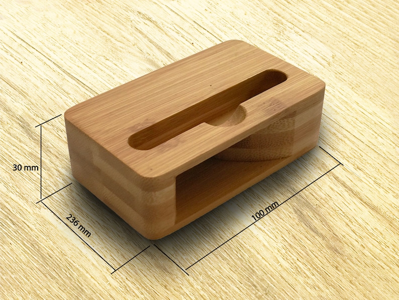 Może przedstawiać: Stojak na telefon z naturalnego bambusa z wycięciem na telefon. Stojak ma wymiary 100 mm x 236 mm x 30 mm.