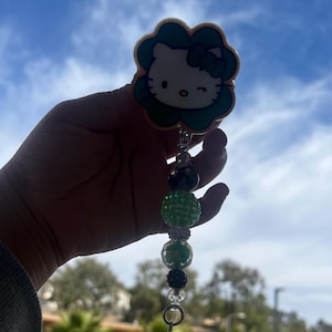 May include: A Hello Kitty retractable badge reel with a green and white design. The badge reel has a flower-shaped border and a beaded charm with green and black beads, and a clear crystal charm.