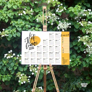 May include: A white poster on a wooden easel, featuring the text "Le Tour de France 2026" with a yellow sun graphic. The poster includes a calendar layout with stage details. The background is a lush green wall with white flowers.