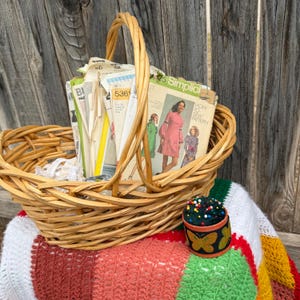 Könnte beinhalten: Ein hellbrauner, geflochtener Weidenkorb gefüllt mit einer Sammlung alter Papierschnittmuster, darunter Umschläge der Marke Simplicity mit Kleiderabbildungen. Daneben steht ein rundes, schwarzes Nadelkissen mit Schmetterlingsmotiven.