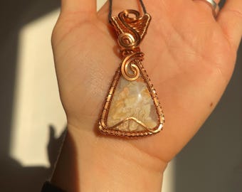 May include: A triangular pendant featuring a polished, cream and tan patterned stone encased in intricate copper wire wrapping. The wire forms decorative swirls at the top and secures the stone along its edges, suspended from a thin black cord.