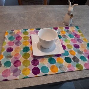 Peut inclure: Une tasse à thé et une soucoupe en céramique blanche sur un set de table à pois colorés. Le set de table a un fond blanc avec des pois roses, violets, bleus, verts et jaunes.