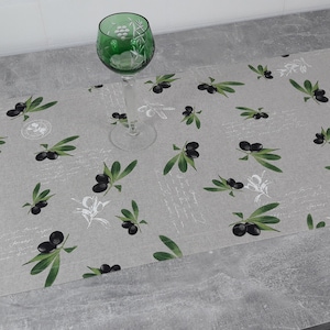May include: A gray linen table runner with a repeating pattern of black olives and green olive branches. A green glass wine glass is on top of the runner.