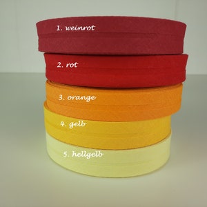 Può includere: Cinque rotoli di nastro in tessuto in varie tonalità di rosso, arancione e giallo. I rotoli sono impilati uno sopra l'altro, con il colore più scuro in basso e il colore più chiaro in alto. Il tessuto è una trama semplice con un leggero luccichio. I rotoli sono etichettati con numeri e parole tedesche per i colori: 1. weinrot, 2. rot, 3. orange, 4. gelb, 5. hellgelb.