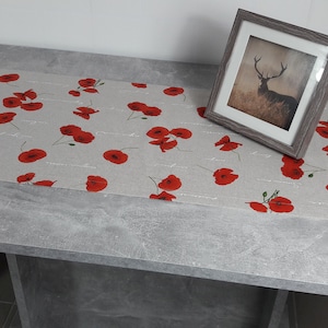 Könnte beinhalten: Ein grauer Tisch mit einem Tischläufer aus Leinen, der mit einem roten Mohnblumenmuster verziert ist. Ein gerahmtes Bild eines Hirsches auf einer Wiese steht auf dem Tisch.
