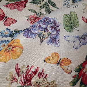 Peut inclure: Un tissu de tapisserie beige avec un motif floral et de papillons. Le tissu présente une variété de fleurs, notamment des fleurs violettes, rouges et jaunes, ainsi que des papillons dans des tons de rouge, jaune et blanc.