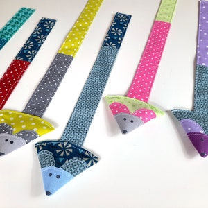 Pode incluir: Seis marcadores de página de tecido em forma de rato com padrões diferentes. Cada marcador de página tem uma cabeça de rato triangular com olhos e orelhas bordados.