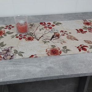 Könnte beinhalten: Ein beiger Tischläufer mit einem floralen Muster mit roten, rosa und grünen Blumen und Vögeln. Der Läufer liegt auf einem grauen Tisch mit einem durchsichtigen Glasgefäß gefüllt mit roten Perlen.