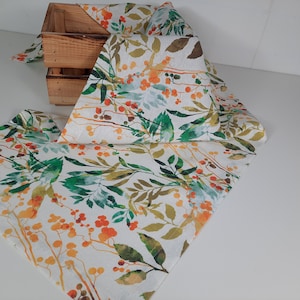 May include: A white linen table runner with a floral pattern featuring green leaves, orange and yellow berries, and a watercolor effect.