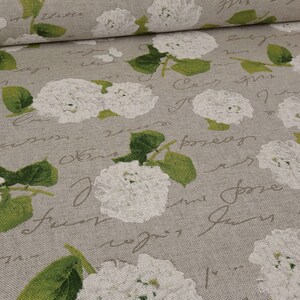 Op de afbeelding: Een beige stof met een herhalend patroon van witte hortensiabloemen, groene bladeren en kleine witte vlinders. De achtergrond is bedekt met een decoratief cursief handschrift in een donkergrijze tint, wat een gelaagd visueel effect geeft.