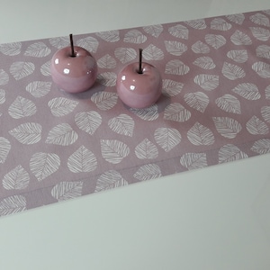 Könnte beinhalten: Ein rosa Tischläufer mit einem weißen Blatt-Muster. Zwei rosa Keramikäpfel liegen auf dem Läufer.