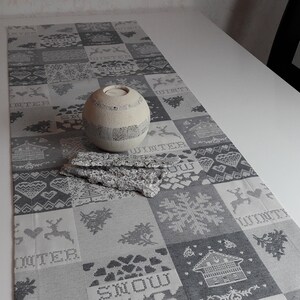 May include: A gray and white table runner with a winter theme. The runner features a variety of patterns, including snowflakes, hearts, and reindeer. The word "Winter" is repeated throughout the runner. A white ceramic candle holder is placed on the runner.