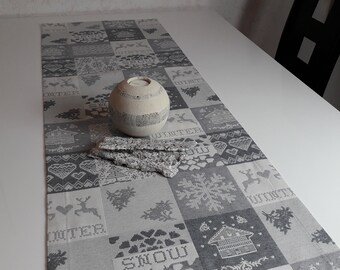 May include: A gray and white table runner with a winter theme. The runner features a variety of patterns, including snowflakes, hearts, and reindeer. The word "Winter" is repeated throughout the runner. A white ceramic candle holder is placed on the runner.