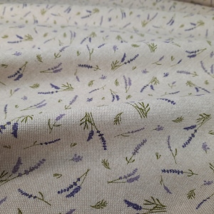 Op de afbeelding: Een close-up van een beige stof met een herhalend patroon van kleine paarse lavendelbloemen en groene stelen.