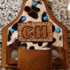 May include: A brown leather belt holder with a leopard print design and turquoise accents. The holder has a metal ring and the initials "CH" are embossed on the front.