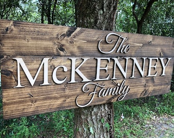 May include: A wooden sign with the text "The McKenney Family" in white raised letters. The sign is stained a dark brown color and has a rustic look.