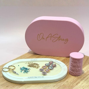 Peut inclure: Une boîte rose ovale avec le texte "On A String" en lettres dorées est derrière un plateau ovale blanc avec des perles, des ciseaux et une boucle d'oreille perlée. Un cylindre rose avec "Size S, 11, 15" est à droite.