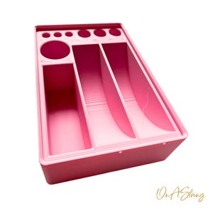 以下が含まれることがあります： 複数のコンパートメントと円形の穴があるピンク色のオーガナイザー。オーガナイザーは長方形で、さまざまなサイズのスロットと大きな円形の開口部があります。「On A String」というテキストが右下にあります。