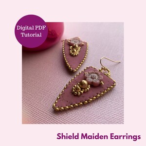 Könnte beinhalten: Rosa-farbene, dreieckige Ohrringe mit goldenen Verzierungen und rosa Blütenakzenten. Die Ohrringe heißen "Shield Maiden Earrings" und sind als digitales PDF-Tutorial erhältlich.