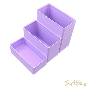 May include: Three rectangular, lavender-colored storage containers of varying sizes are stacked in a stepped arrangement. The containers are empty and have a smooth, matte finish. The arrangement is set against a white background.