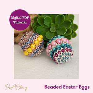 Może przedstawiać: Dwa koralikowe jajka wielkanocne, jedno z żółtymi, różowymi i niebieskimi koralikami, a drugie z różowymi, turkusowymi i srebrnymi koralikami. Jajka leżą na zielonej roślinie sukulentowej.