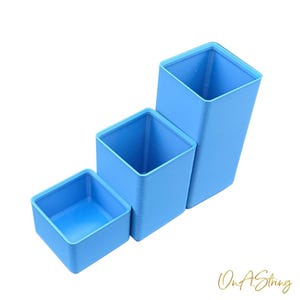 May include: Three light blue square containers of varying sizes are stacked. The containers have a smooth, matte finish and are empty. The containers are arranged in a stepped pattern, with the smallest container in the front.