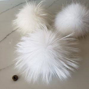 May include: Close-up of three fluffy white pom-poms with long, soft, feathery strands. One pom-pom is a light cream color. A small, round, bronze-colored snap is also visible. These are likely for hats or other crafts.