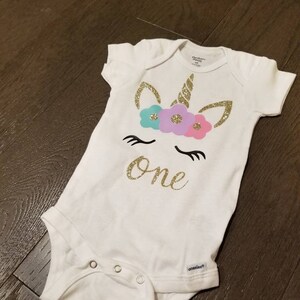 May include: White onesie with a gold glitter unicorn with a flower crown and the word "one" in gold glitter.