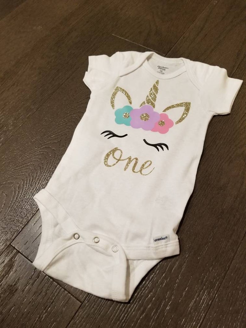 May include: White onesie with a gold glitter unicorn with a flower crown and the word "one" in gold glitter.