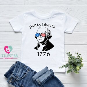 May include: White t-shirt featuring a black and white graphic of George Washington wearing sunglasses with American flag designs. The text "party like its 1776" is above the image. Blue jeans are folded below the shirt.