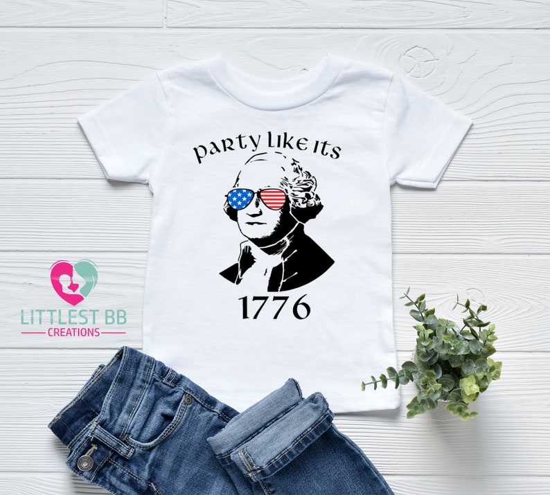 May include: White t-shirt featuring a black and white graphic of George Washington wearing sunglasses with American flag designs. The text "party like its 1776" is above the image. Blue jeans are folded below the shirt.