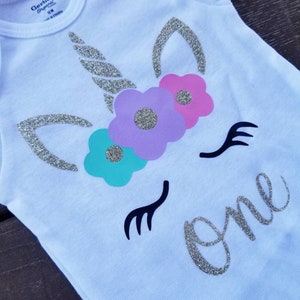 May include: White onesie with a silver glitter unicorn design and the word "one" in silver glitter. The unicorn has a silver glitter horn, purple, pink, and teal flowers, and black eyelashes.
