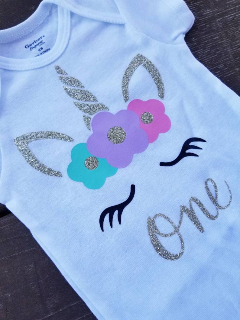 May include: White onesie with a silver glitter unicorn design and the word "one" in silver glitter. The unicorn has a silver glitter horn, purple, pink, and teal flowers, and black eyelashes.