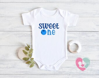 May include: White baby onesie with blue text that reads "Sweet One" and a blue blueberry graphic.