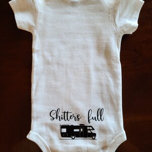 Op de afbeelding: Witte baby romper met zwarte tekst die "Shitters full" zegt en een zwart silhouet van een recreatievoertuig.