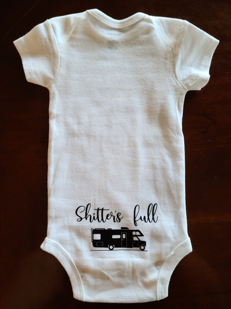 Op de afbeelding: Witte baby romper met zwarte tekst die "Shitters full" zegt en een zwart silhouet van een recreatievoertuig.